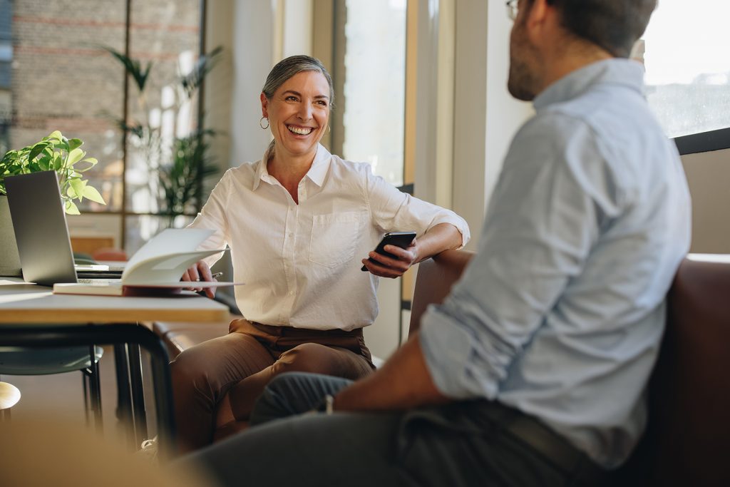 A woman sitting at a desk with a laptop on it chatting to a man sitting across from her