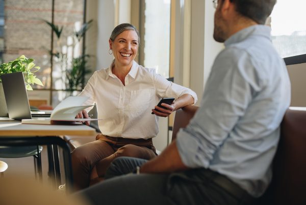 A woman sitting at a desk with a laptop on it chatting to a man sitting across from her