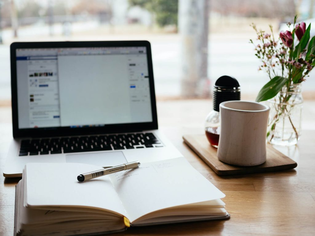 A desk with a laptop open to a blank document with a notebook open in front of it. Next to it are a pen, coffee cup and flowers