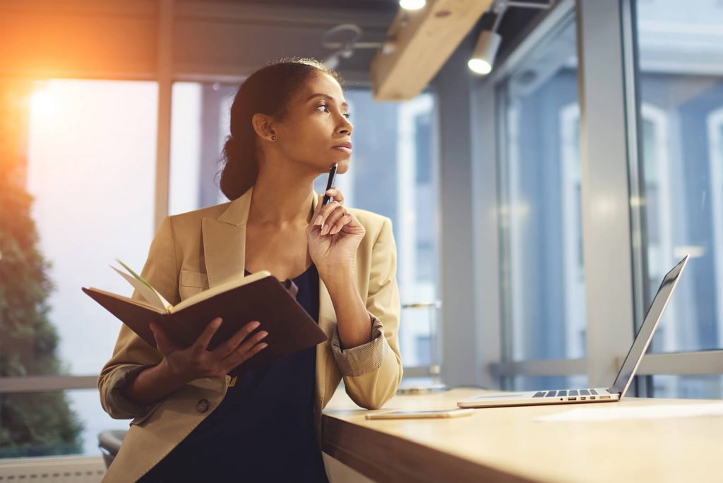 woman leaning against a desk looking out the window with a pen and notebook in hand