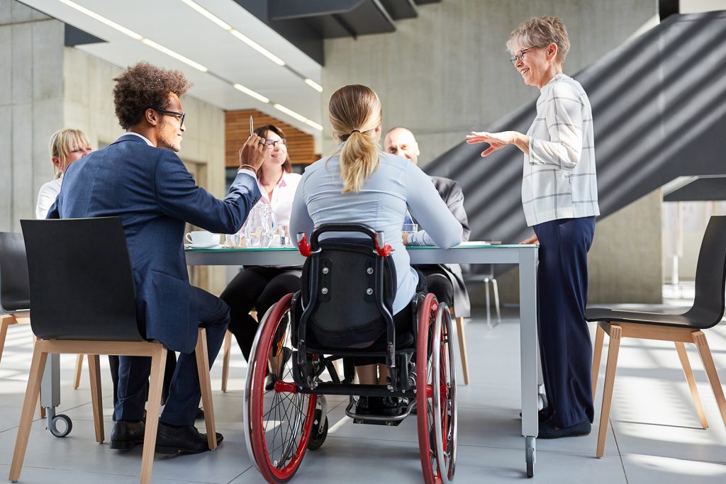 Group of 6 business people sitting around a table discussing, one of the individuals is in a wheelchair