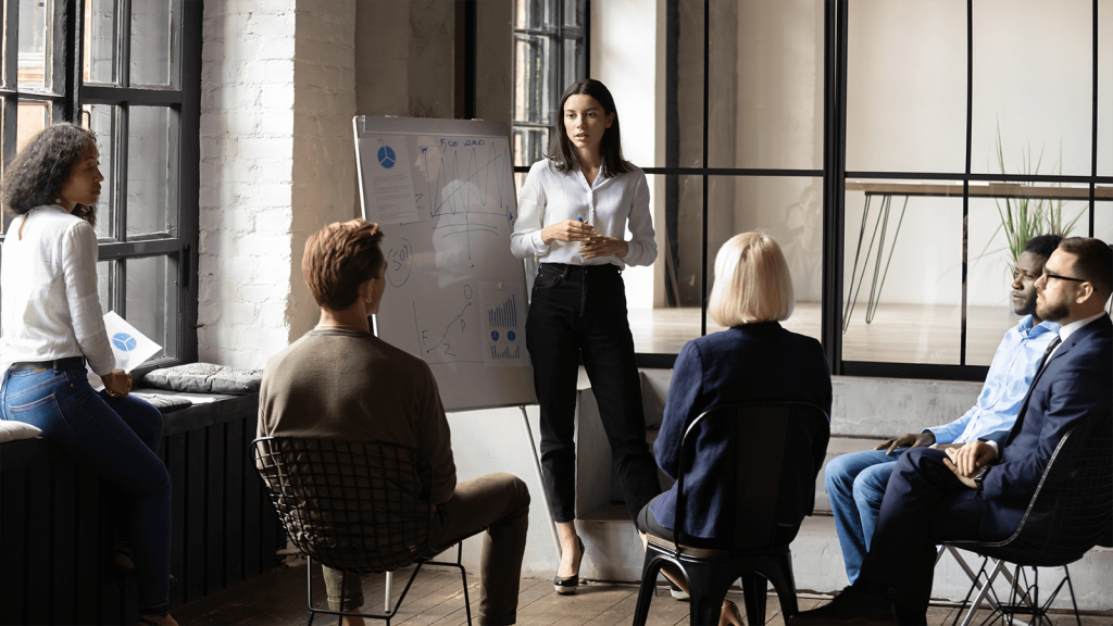 A woman standing next to a whiteboard speaking to a group of seated people