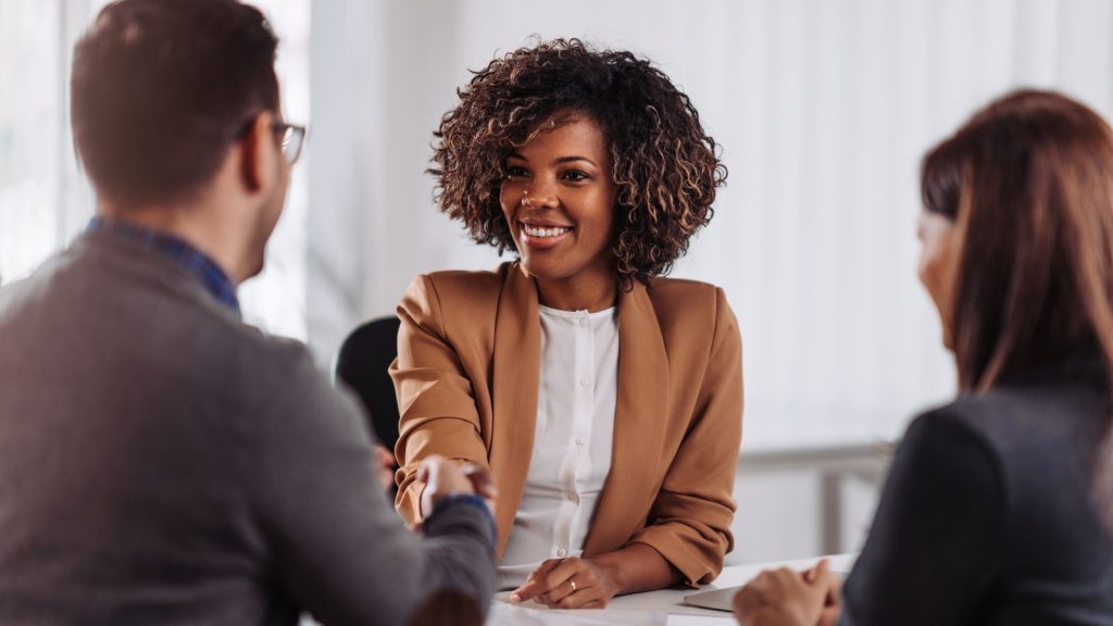 Thee individuals sitting at a table wearing business atire. A woman at the table is shaking a man's hand.