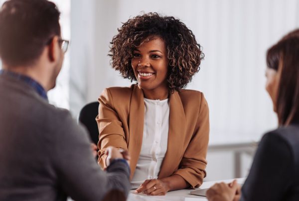 Thee individuals sitting at a table wearing business atire. A woman at the table is shaking a man's hand.