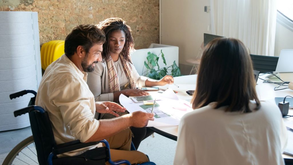 Three individuals in business attire sitting at a desk looking at work documents. One of the individuals in sitting in a wheelchair at the table, the two others are sitting on chairs.