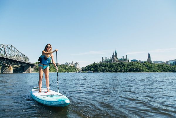 A woman standing on a blue and white paddleboard rowing while on a river. There is Ottawa Parliament building and bridge in the background.