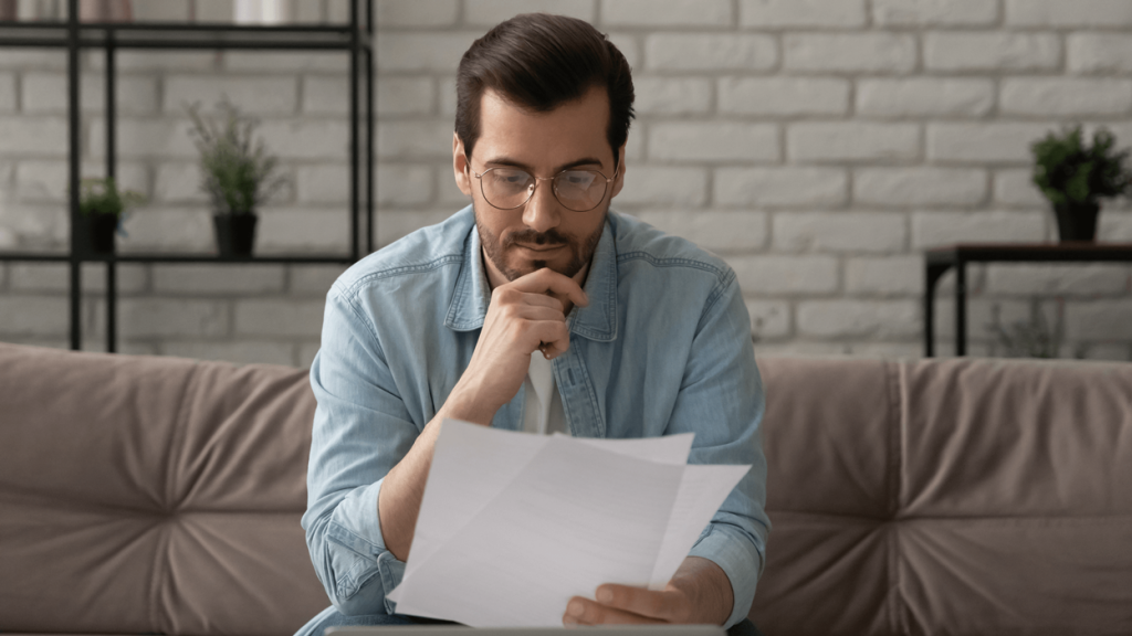 A man sitting on a couch looking at documents that are in one hand while his other hand rests on his chin pensively