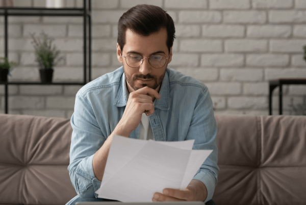 A man sitting on a couch looking at documents that are in one hand while his other hand rests on his chin pensively