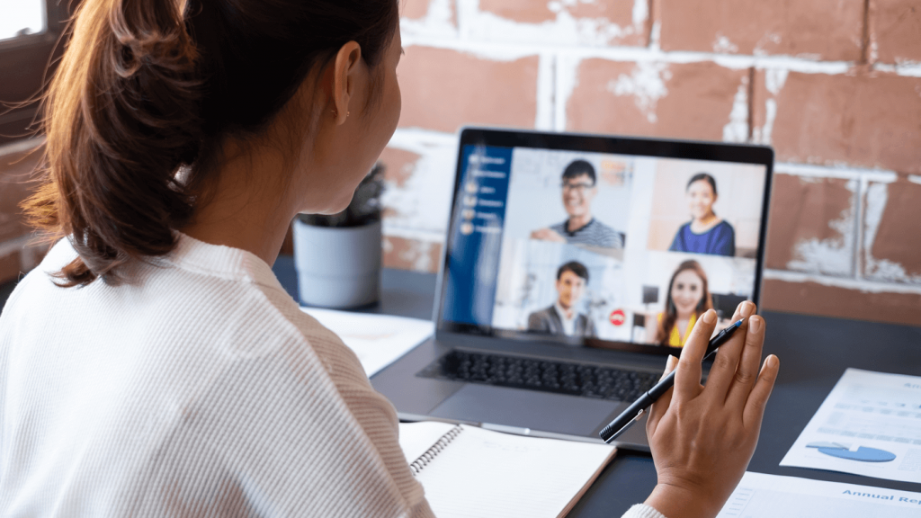 A woman sitting at a desk video chatting off a laptop with four other individuals in professional attire on the screen.