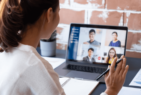 A woman sitting at a desk video chatting off a laptop with four other individuals in professional attire on the screen.
