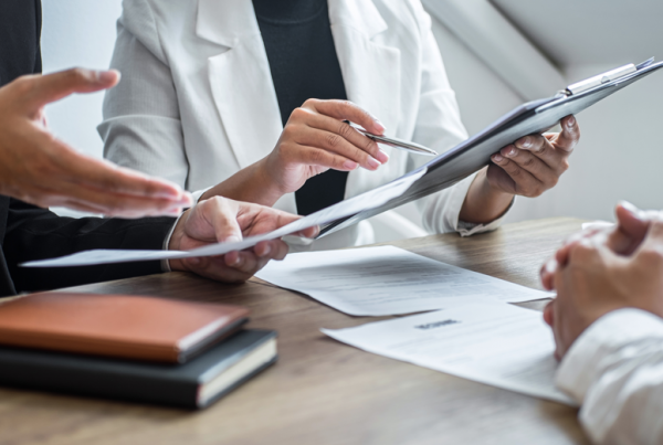 There 3 people sitting at a desk wearing business attire. You only see the individuals neck down. 2 Individuals are on one side holding papers in their hands, the other is on the other side of the desk with his hands crossed.