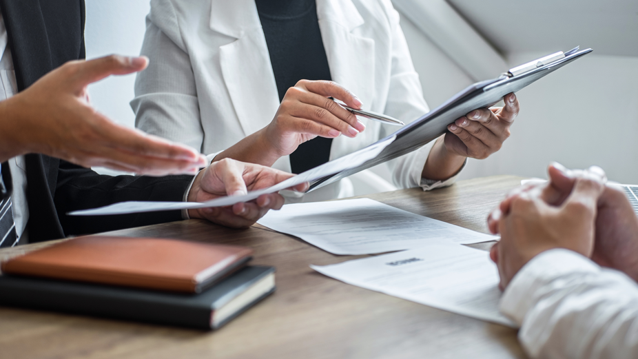 There 3 people sitting at a desk wearing business attire. You only see the individuals neck down. 2 Individuals are on one side holding papers in their hands, the other is on the other side of the desk with his hands crossed.