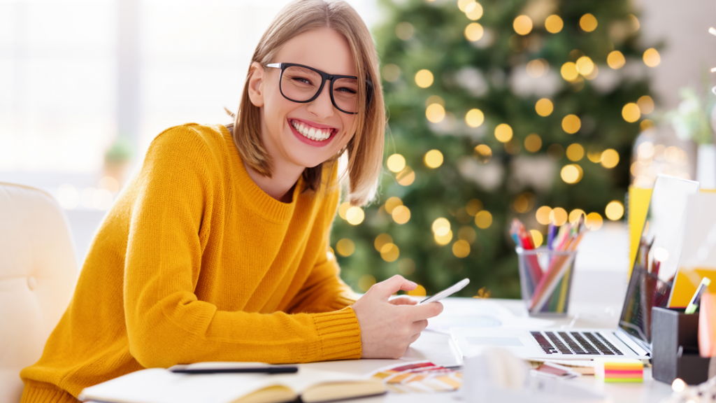 A woman smiling while sitting at a desk with a laptop and notes on it. In the background there is a Christmas tree.