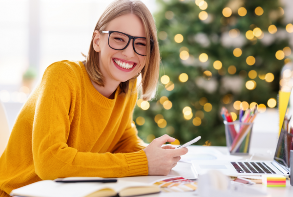 A woman smiling while sitting at a desk with a laptop and notes on it. In the background there is a Christmas tree.