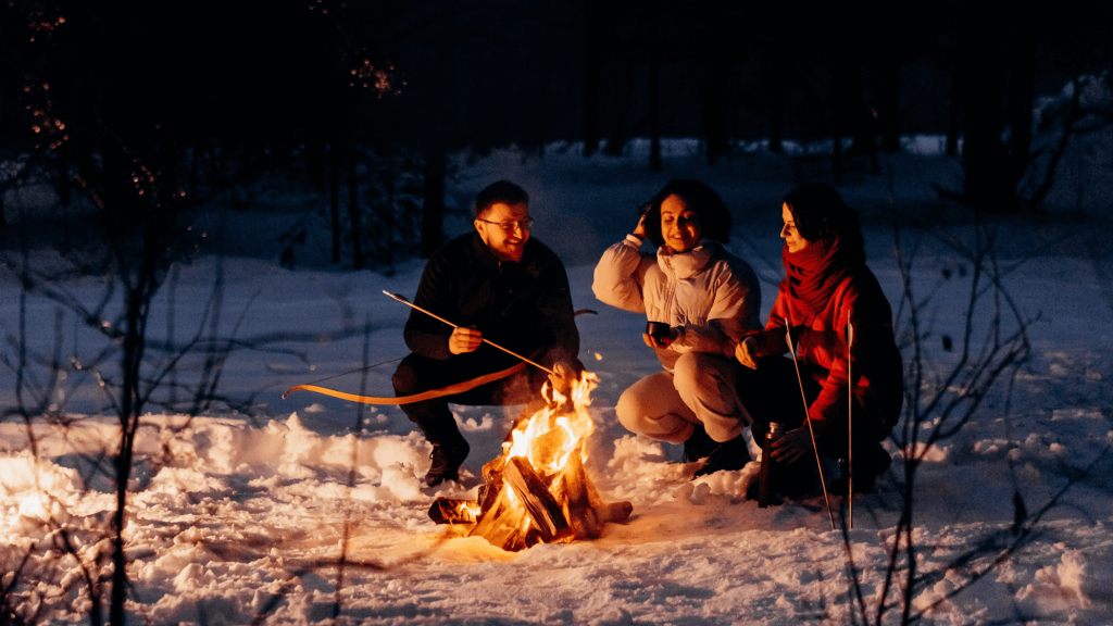 Three individuals in snowsuits sitting around an outdoor fire in the forest with snow on the ground.