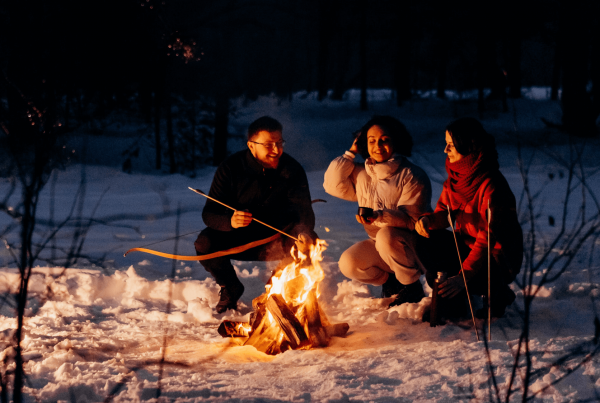 Three individuals in snowsuits sitting around an outdoor fire in the forest with snow on the ground.