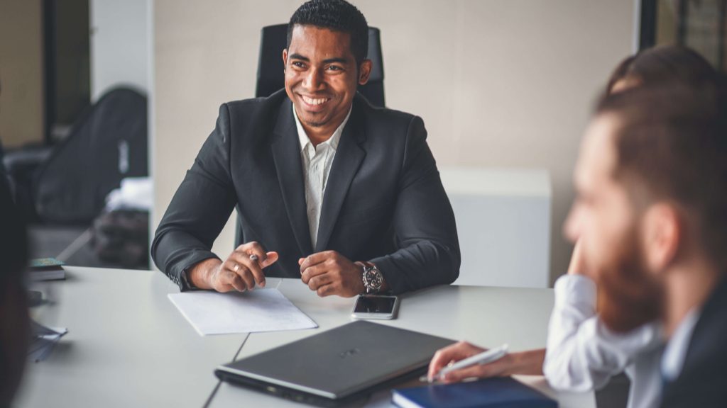 A man in a suit smiling while sitting at a table across from 3 other individuals in business attire. There are papers and a laptop on the table. In the background we see an office.