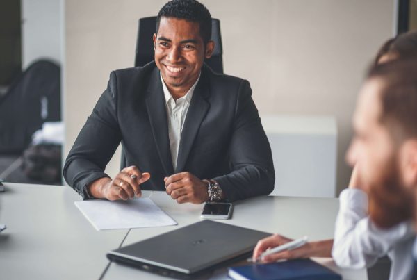 A man in a suit smiling while sitting at a table across from 3 other individuals in business attire. There are papers and a laptop on the table. In the background we see an office.