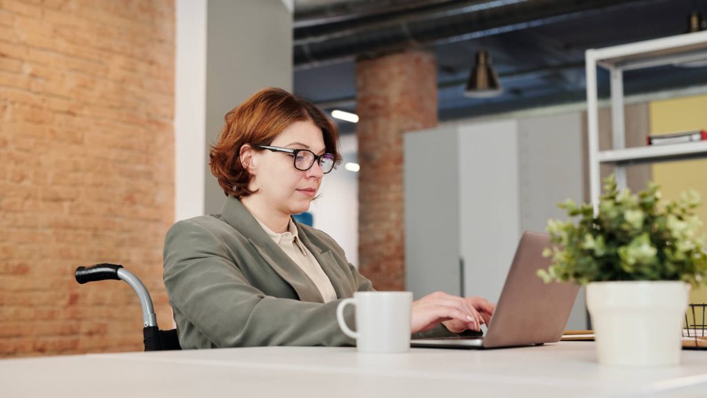 A woman sitting in a wheelchair at a desk staring intently at a laptop while typing. She is wearing business attire.