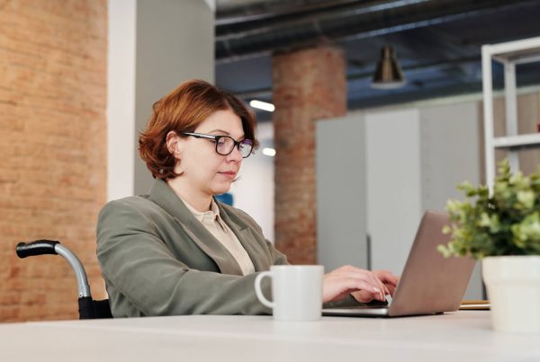 A woman sitting in a wheelchair at a desk staring intently at a laptop while typing. She is wearing business attire.