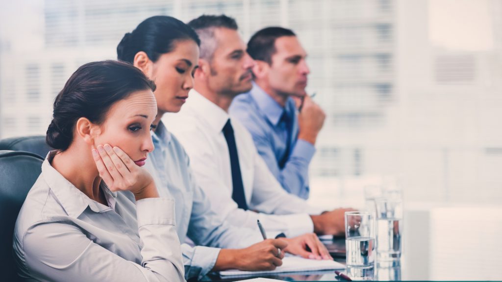 Four individuals in professional attire sitting side by side at a table in an office. The individual closest to the frame as her face resting on her hand with an expression that appears to be disinterested.