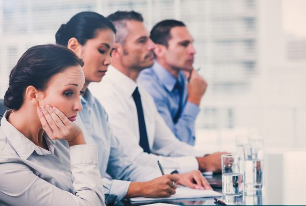 Four individuals in professional attire sitting side by side at a table in an office. The individual closest to the frame as her face resting on her hand with an expression that appears to be disinterested.