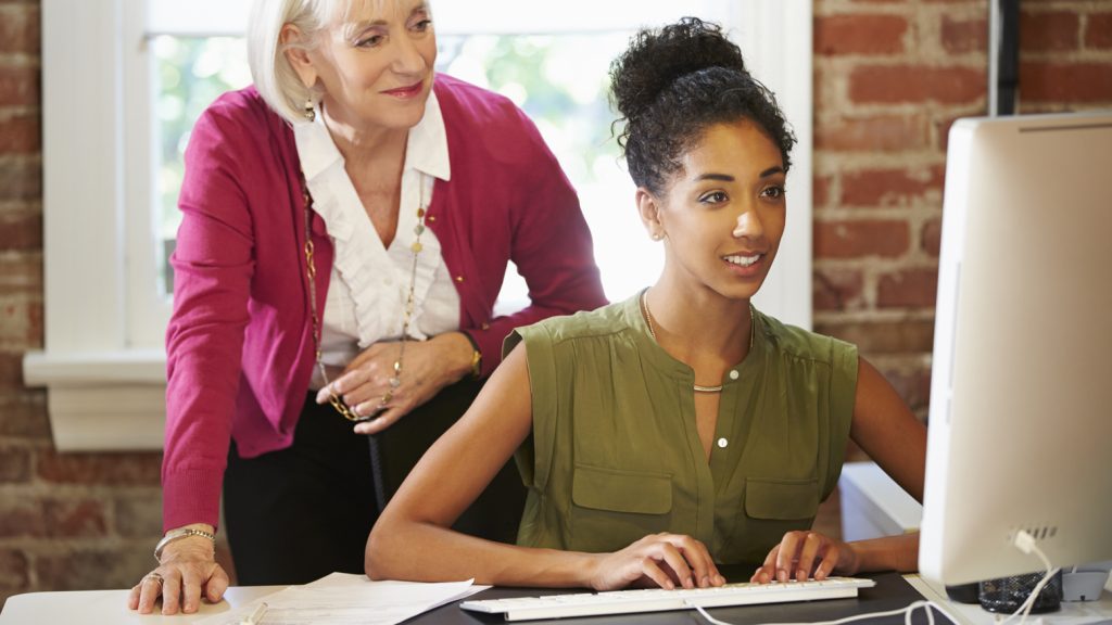 A woman in her 20s sitting at a desk typing on a computer with a woman in her 70s standing behind her looking at the computer screen. Both women are dressed in professional attire.