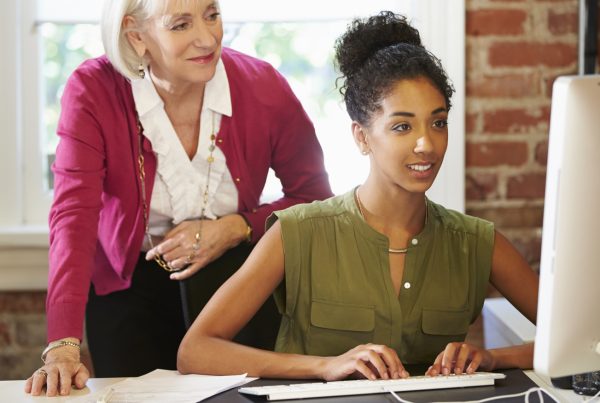 A woman in her 20s sitting at a desk typing on a computer with a woman in her 70s standing behind her looking at the computer screen. Both women are dressed in professional attire.