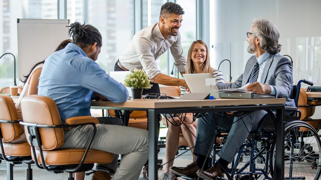 A group of 4 business professionals from diverse background sitting at a table talking. 1 of the individuals is in a wheelchair.