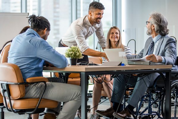 A group of 4 business professionals from diverse background sitting at a table talking. 1 of the individuals is in a wheelchair.