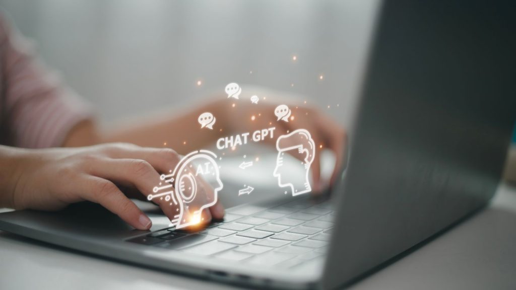 Hands typing on a laptop keyboard with an illustration of a human head and a robot head and the words 'CHAT GPT' in between them appearing above the keyboard.
