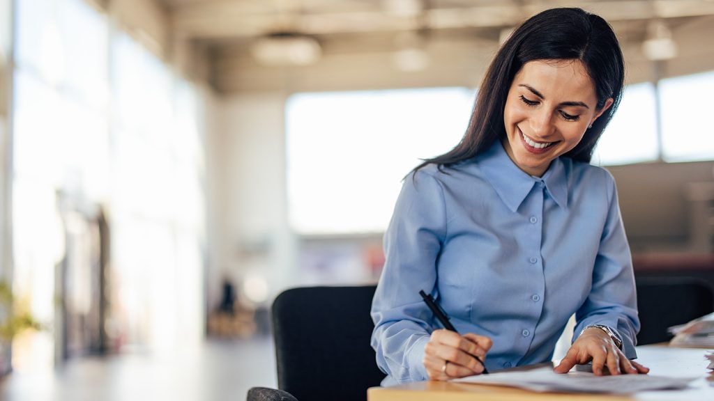 A woman with dark brown hair wearing a blue blouse sitting at a desk writing on a piece of paper. The background is an office setting.