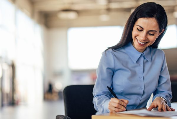 A woman with dark brown hair wearing a blue blouse sitting at a desk writing on a piece of paper. The background is an office setting.