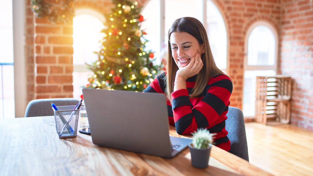 A laptop is on a table with a woman sitting across from it staring at the screen. In the background is a Christmas tree.