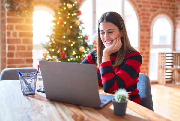 A laptop is on a table with a woman sitting across from it staring at the screen. In the background is a Christmas tree.