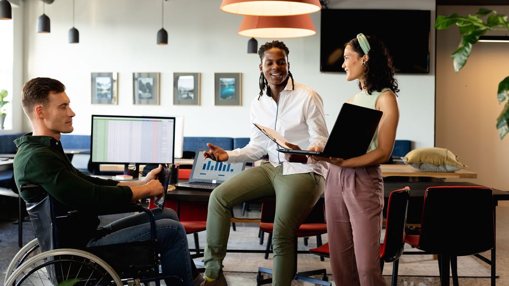 Three individuals around a desk in an office setting chatting. One individual is standing holding a laptop, another is sitting on a desk talking, while the other is sitting in a wheelchair listening.