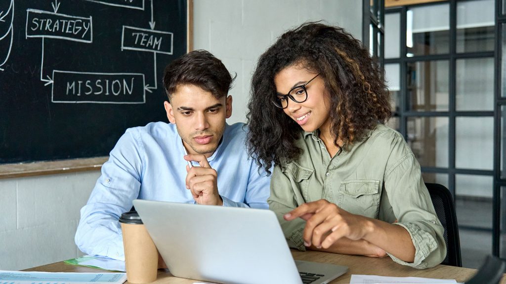 Two young professionals sitting at a desk looking at a laptop screen.