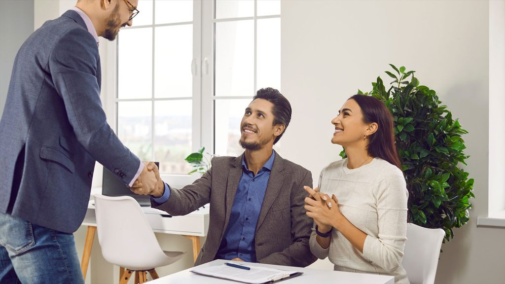 A woman and a man wearing professional clothing sitting down at a desk looking up at a man standing on the other side of the desk. The two men are shaking hands.