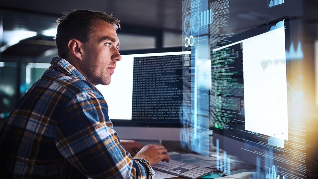 man sitting at a desk looking at a monitor with coding on it