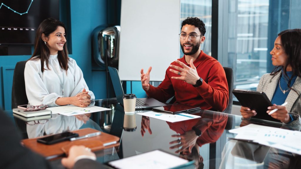 3 individuals in professional clothing sitting at a desk talk. There are papers on the desk.