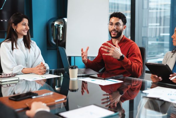 3 individuals in professional clothing sitting at a desk talk. There are papers on the desk.