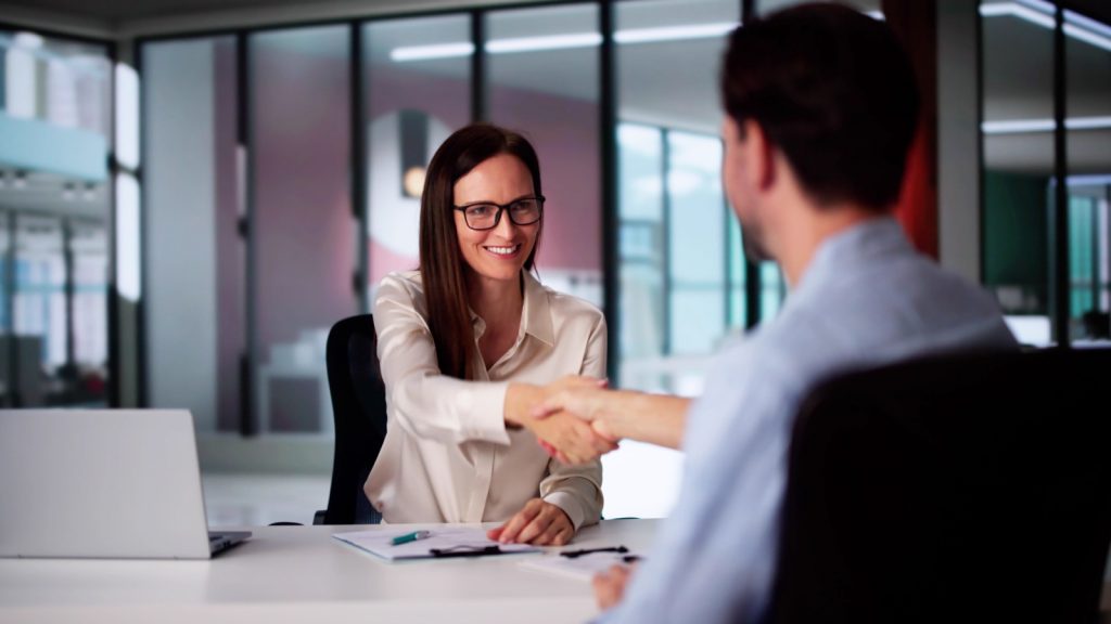 a woman in professional attire sitting at a desk with a laptop shaking the hands with a man sitting across from her