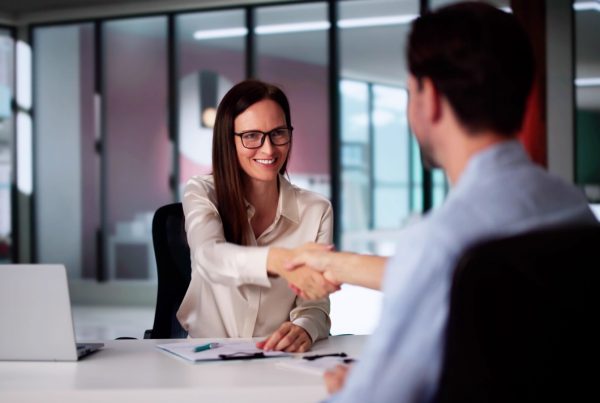 a woman in professional attire sitting at a desk with a laptop shaking the hands with a man sitting across from her