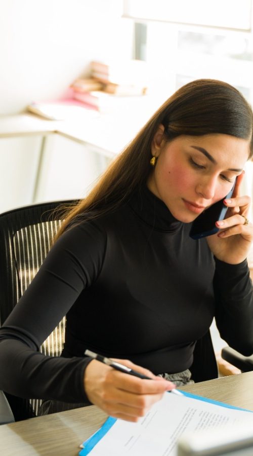 A woman in professional attire sitting at a desk writing notes on a paper while talking on the phone.