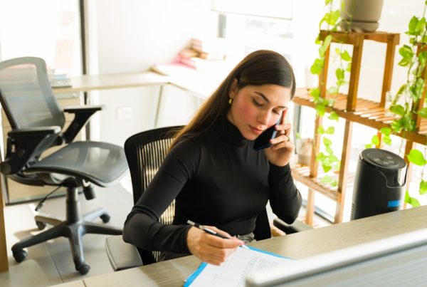 A woman in professional attire sitting at a desk writing notes on a paper while talking on the phone.