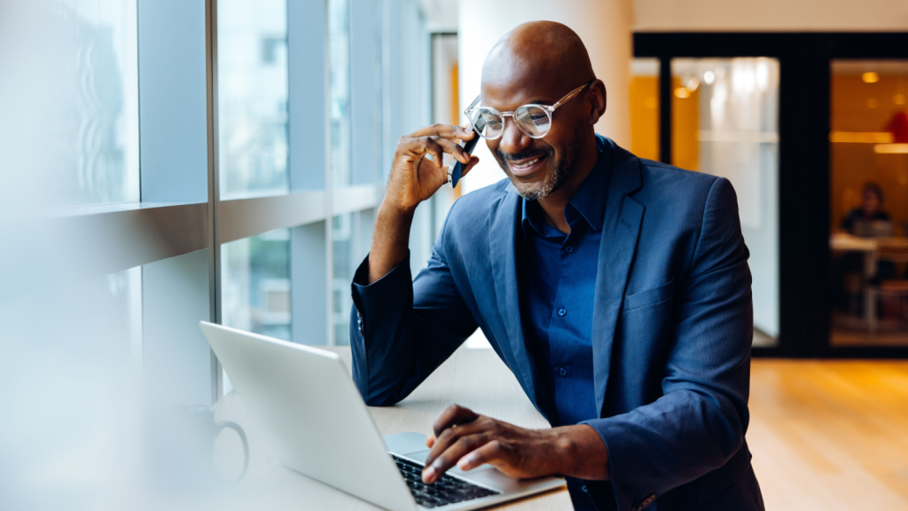 a man speaking on a cellphone while sitting at a desk typing on a laptop