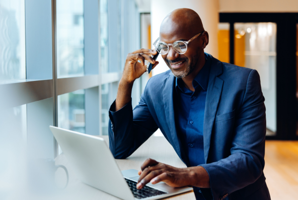a man speaking on a cellphone while sitting at a desk typing on a laptop