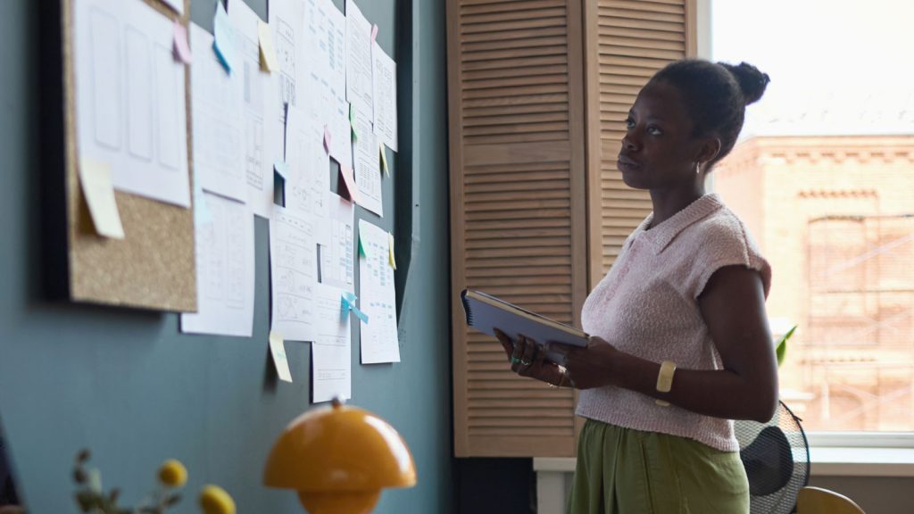 A woman in professional attire standing in her office looking at a corkboard filled with papers
