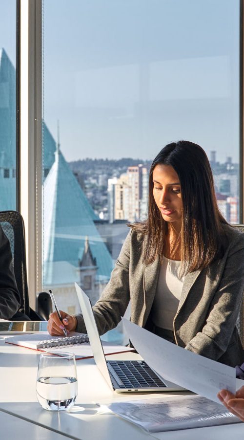 Several individuals wearing professional attire sitting in a boardroom. Some are talking, some are looking at papers.