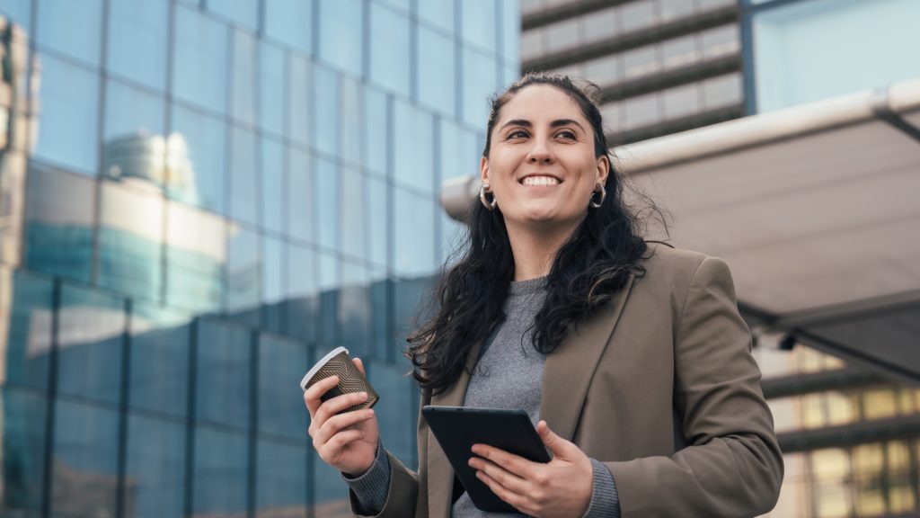 A woman in professional attire standing outside of an office building holding a tablet while smiling